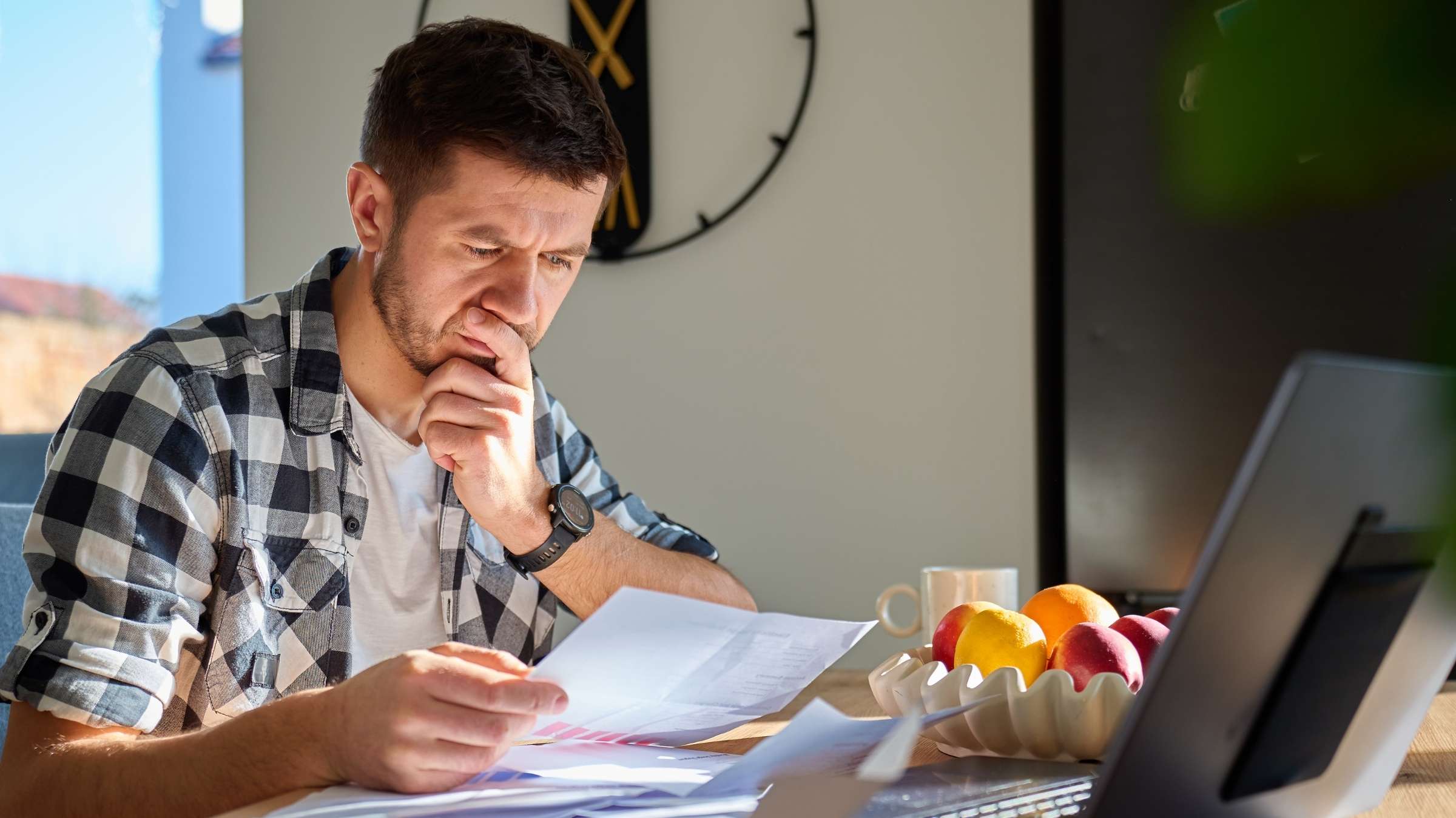 Man in a plaid shirt reads and analyzes documents at a desk with a laptop and a bowl of fruit nearby.